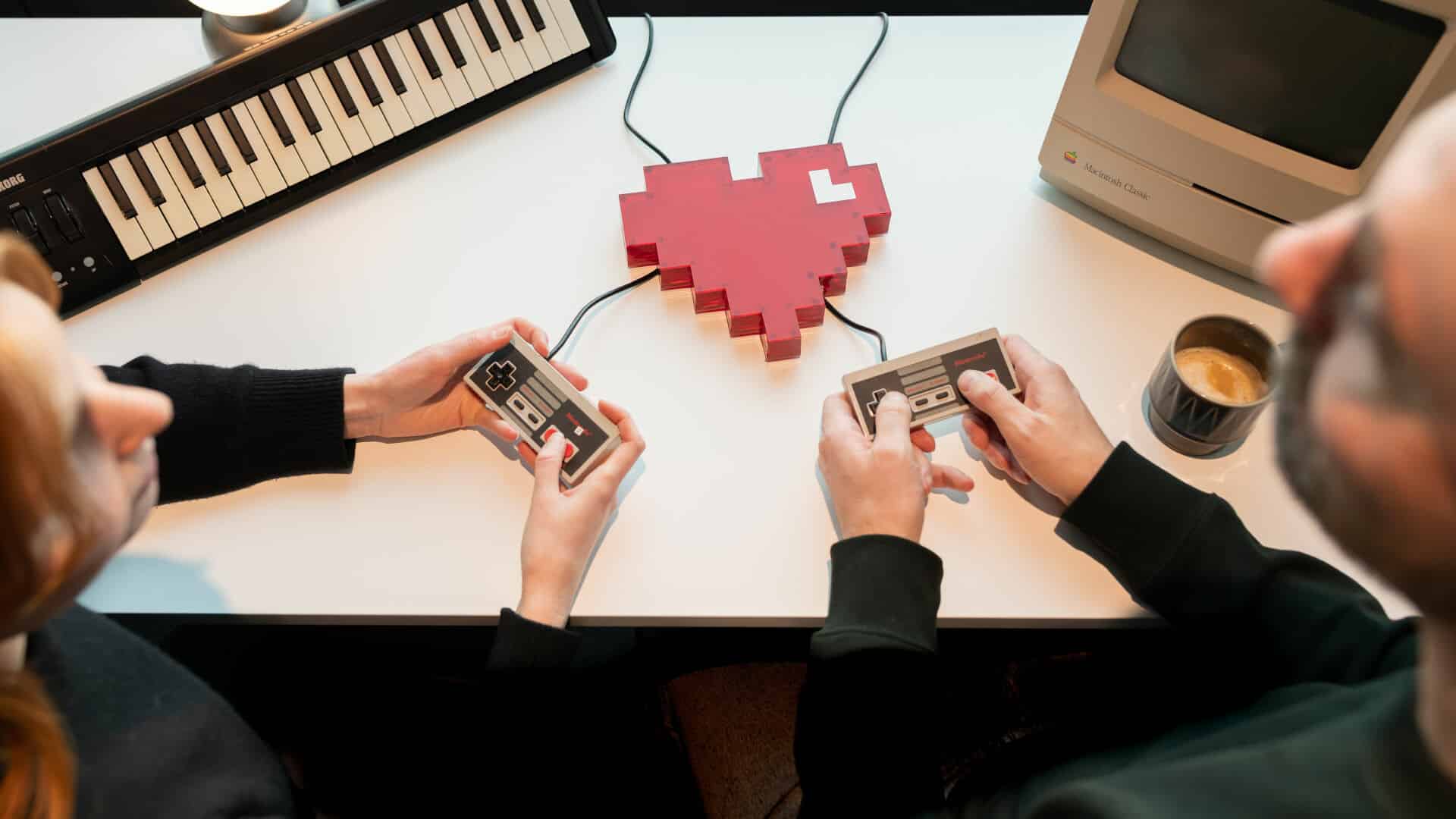 Två personer håller var sin spelkontroll i handen. Mellan dem ligger ett hjärta på ett bord.